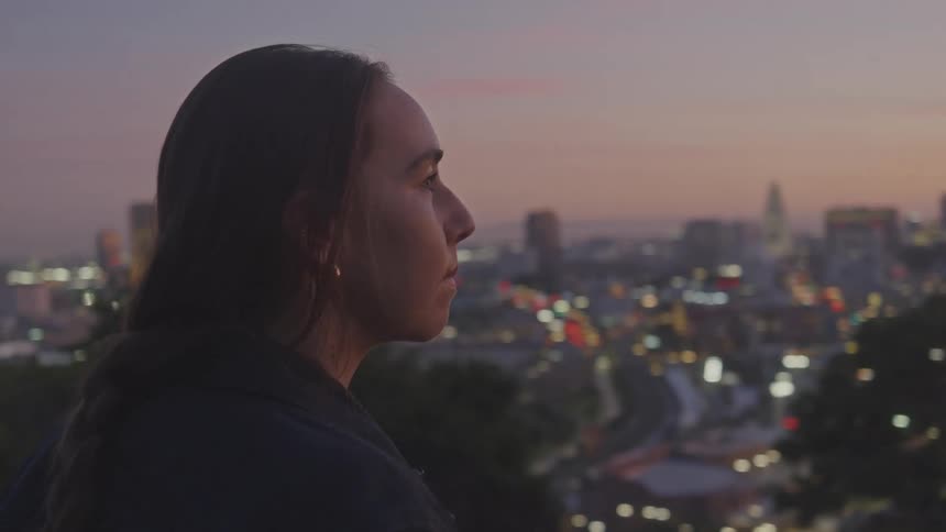 Woman leaning her motorcycle against a hill enjoying the sunset view of a metropolis with skyscrapers