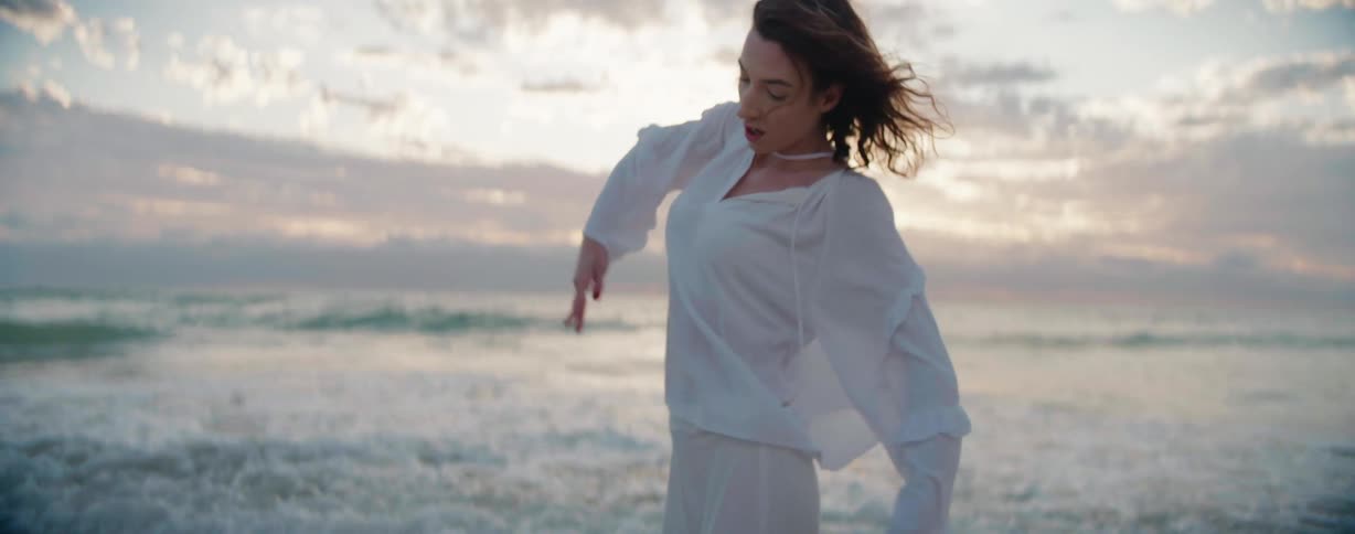 A female dancer with wavy hair a white shirt and pants dances on the beach near the sea at sunset