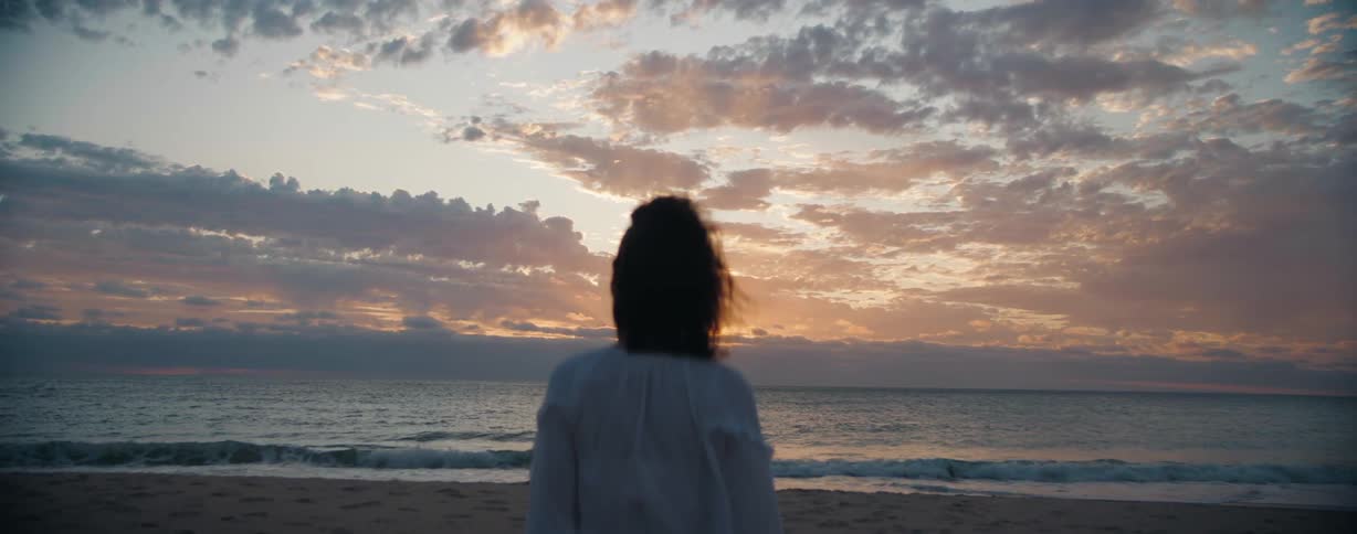 A female facing the ocean while standing on a beach moving her hair at dusk