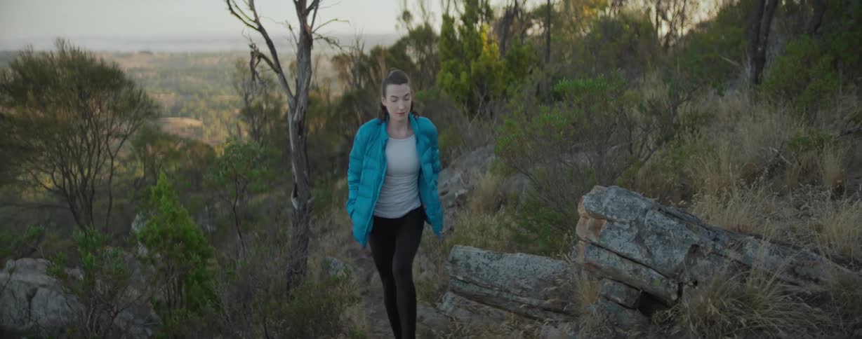 A woman in a blue jacket is walking in a region of nature with a grass and rocks and trees