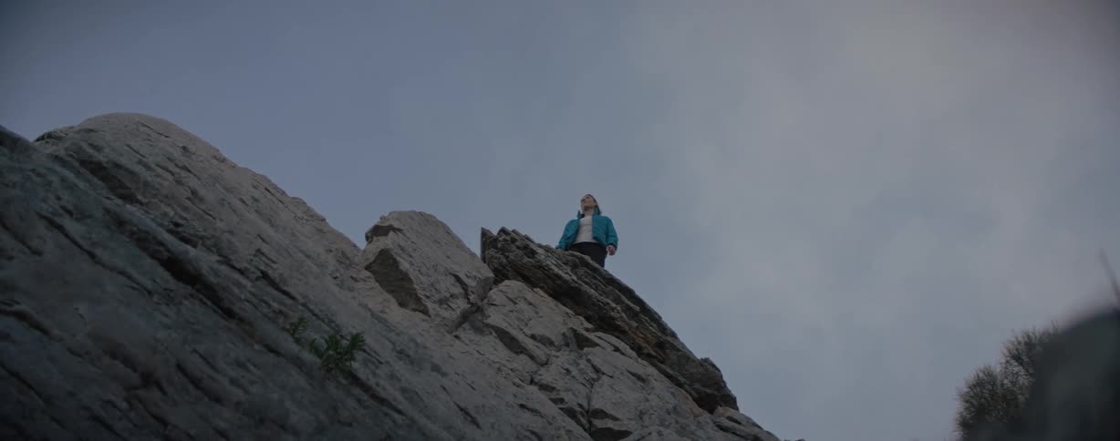 A woman in a blue jacket standing on a high rock meet the blue sky with white clouds passing by