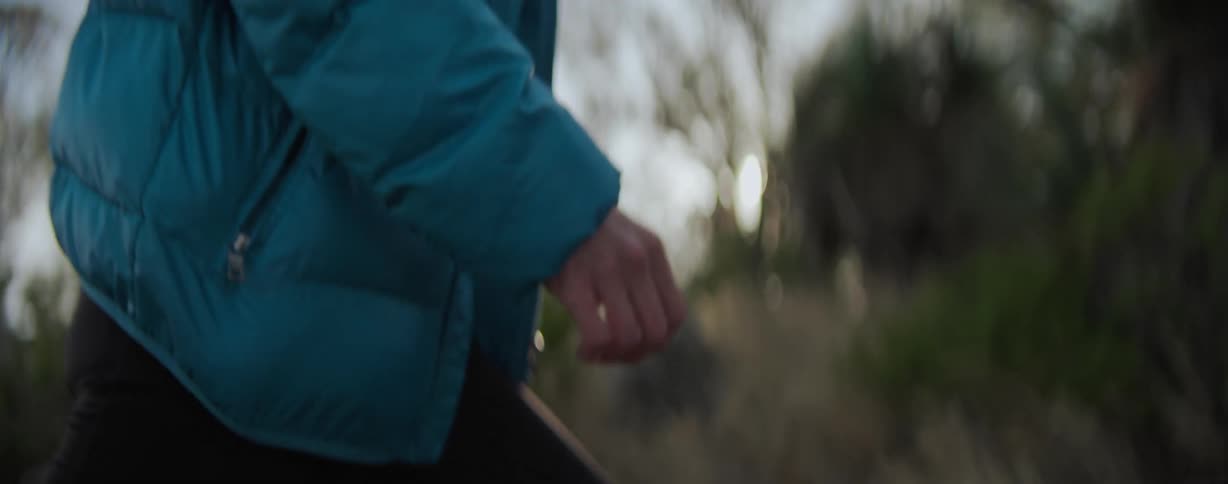 A woman wearing a blue jacket and black pants walks up a mountain slope at sunset