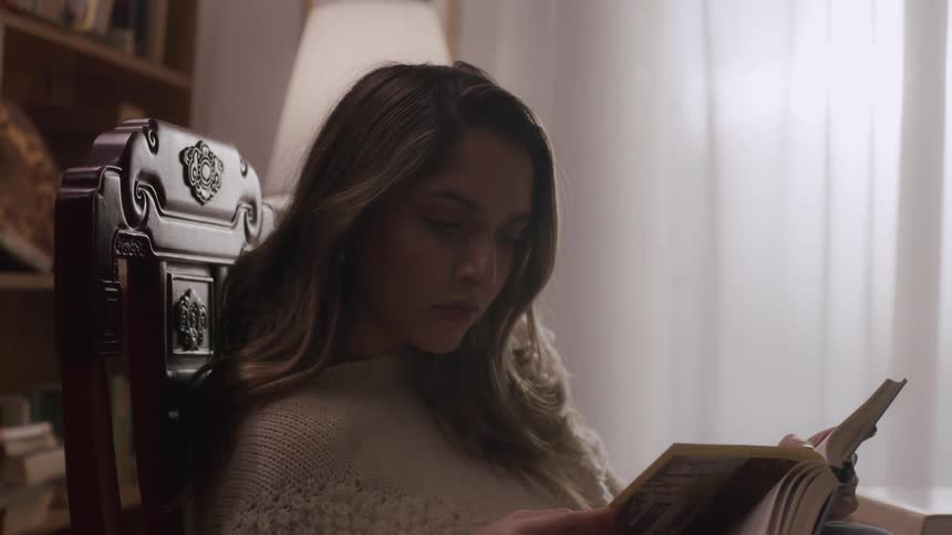 A young woman sits in a chair in a home library and reads a book