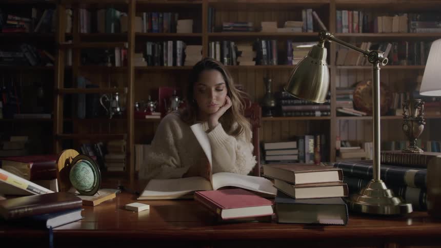 Woman sits down at table near a bookcase and starts reading a book