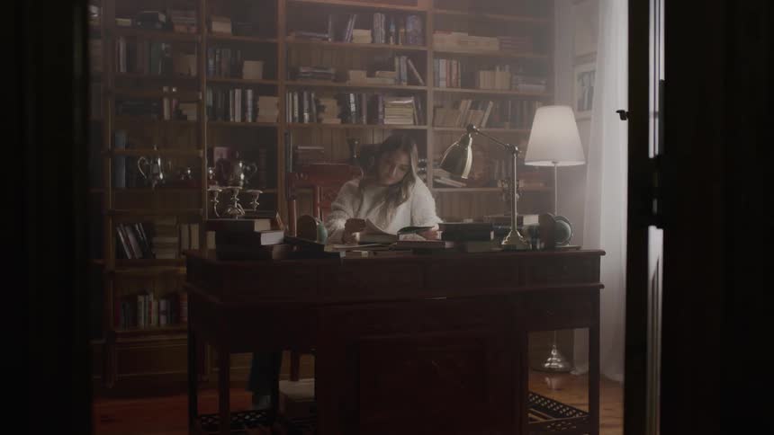 Woman sits at a wooden desk near several bookcases and reads a book