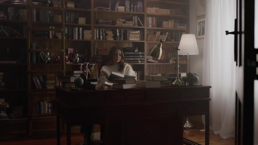 A young woman pulls a book from a shelf sits down at a desk and begins reading