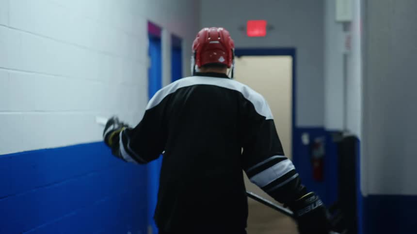 A hockey player wearing full gear picks up a stick and walks down a hallway