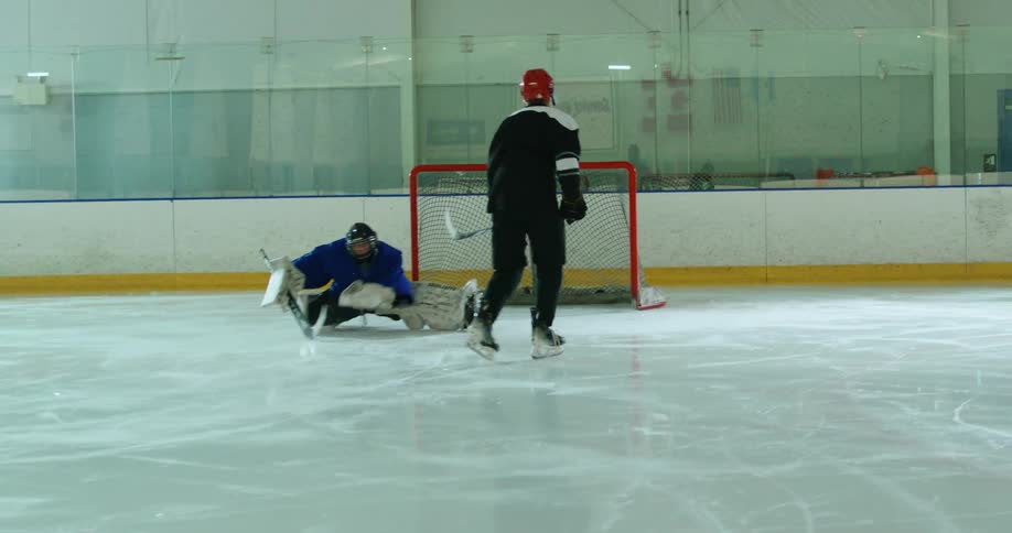 Hockey player with helmet and stick throwing a puck towards the goal against a goalkeeper