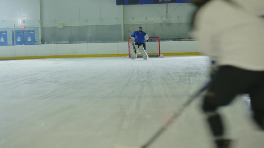 Two individuals on an ice rink with a goalie in blue clothing and a woman in white with a hockey stick scoring a point