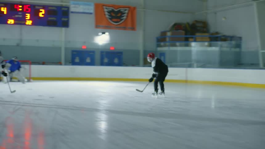 Two players skate towards the goalie and shoot a puck on an ice hockey rink
