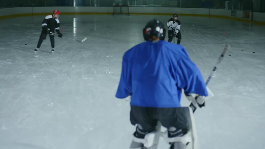 A hockey goalkeeper is wearing blue clothing and is going to defend the attack of two opposing players who are coming in front of him