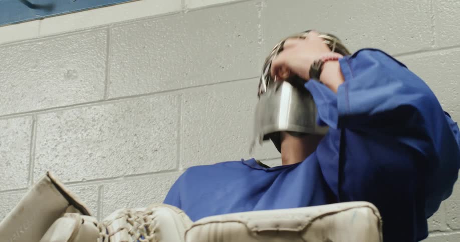 A teenager wearing hockey gear sits on a bench and takes out his helmet