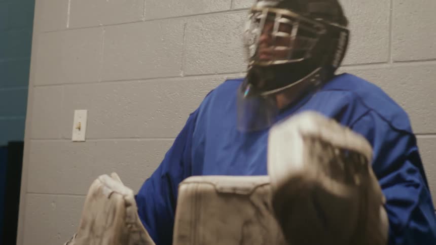 A male hockey goalie sits on a wooden bench and takes off his helmet