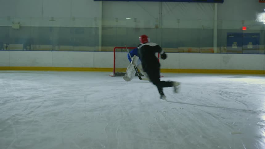 A hockey player in a black jersey and red helmet skates towards the goal preparing to take a shot while the goalie in blue gear braces for the save