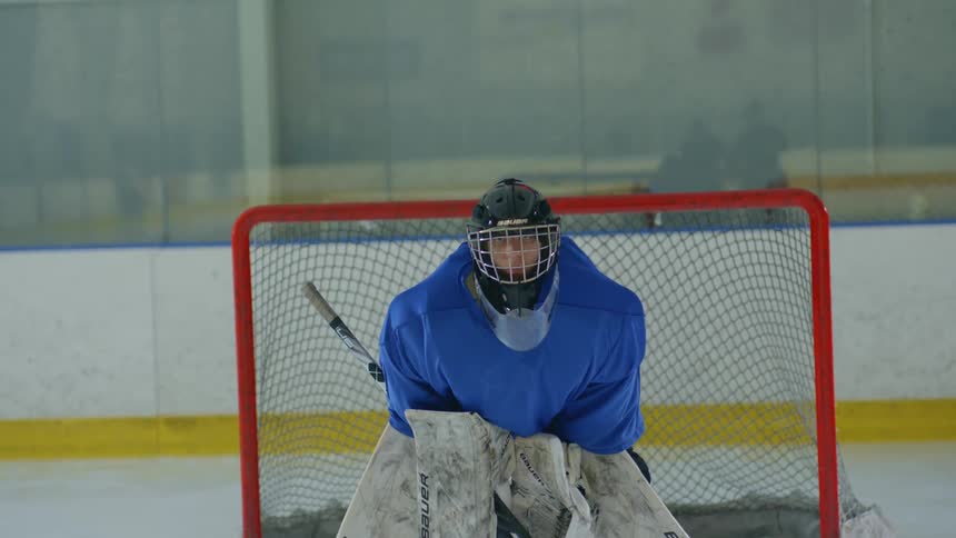 The goalie fully geared up in blue and white equipment stands alert in front of the hockey net ready to defend against an incoming shot