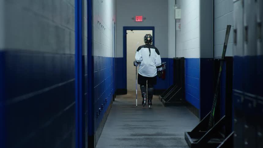 A hockey player wearing a helmet and a white jersey picks up a hockey stick and walks out of the hall heading towards the ice rink