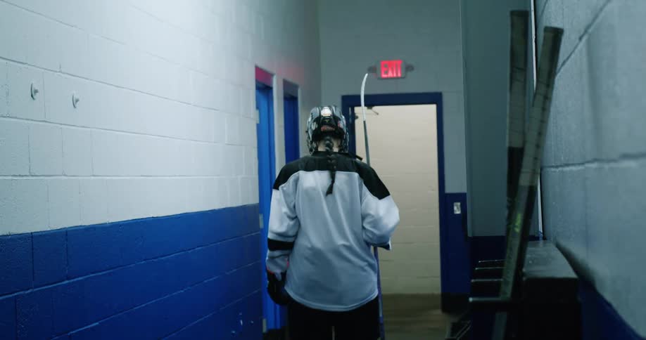 A hockey player wearing a helmet and a white jersey picks up a hockey stick and walks out of the hall heading towards the ice rink