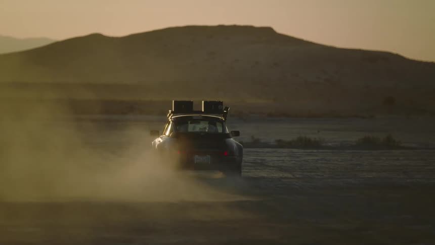 A car is driving through the desert with mountains in the background