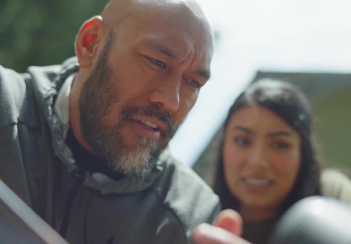 A tan, bald man with a beard looks down at something and explains it to a dark-haired, young woman who is looking down over his shoulder with a questioning look on her face.