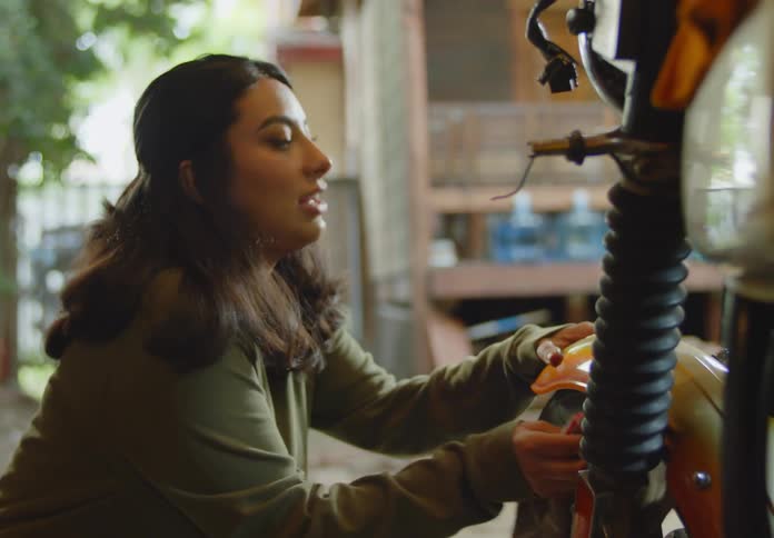 A young dark-haired woman squatting down in a workshop sprays a rag and uses it to clean the wheel of an orange motorbike while she chats with someone in the room.