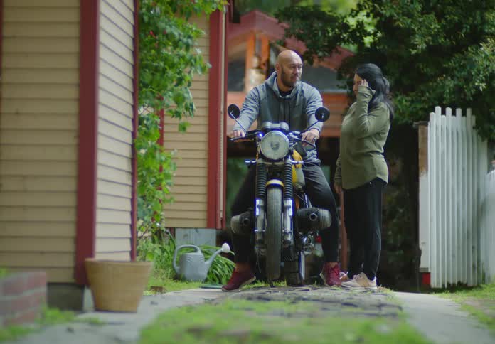 A bald, bearded man and a dark-haired woman laugh together in their front driveway while discussing and testing the handlebars of the motorcycle he is sitting on as  their dogs walk around in the foreground.