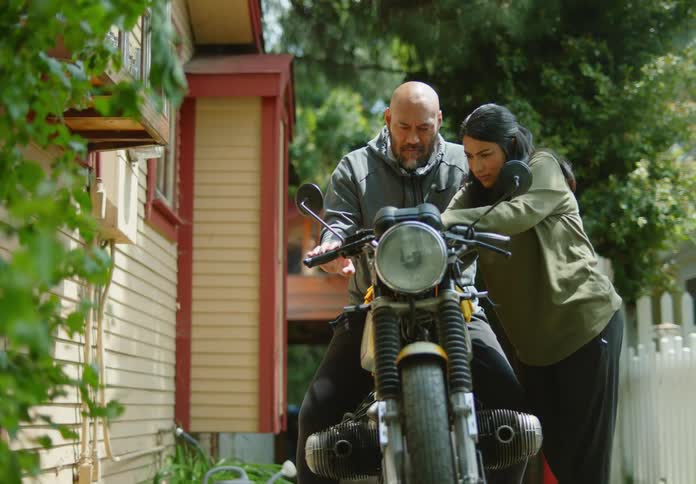 A bald, bearded man sits on a motorcycle and interacts with a dark-haired young woman as they fiddle with the buttons, gears, and handlebars in a rural driveway.