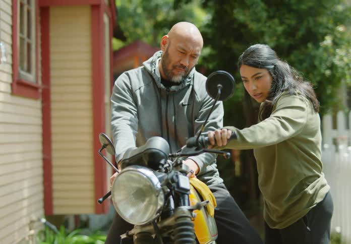 A bald, bearded man is showing off his motorcycle in a driveway to a dark-haired young woman who checks it out while they smile, laugh and flirt.