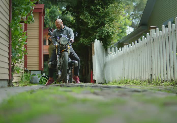 A bald, bearded man in a hoodie is sitting on a parked motorcycle while talking with a dark-haired woman who is walking around examining it