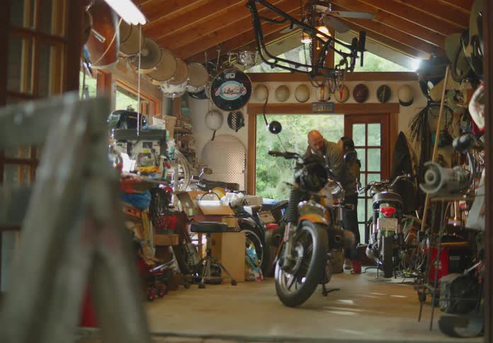 A man and a woman walk into a cluttered garage workshop with boxes in hand and then put them down as they walk through taking a look at the motorcycles and other things inside