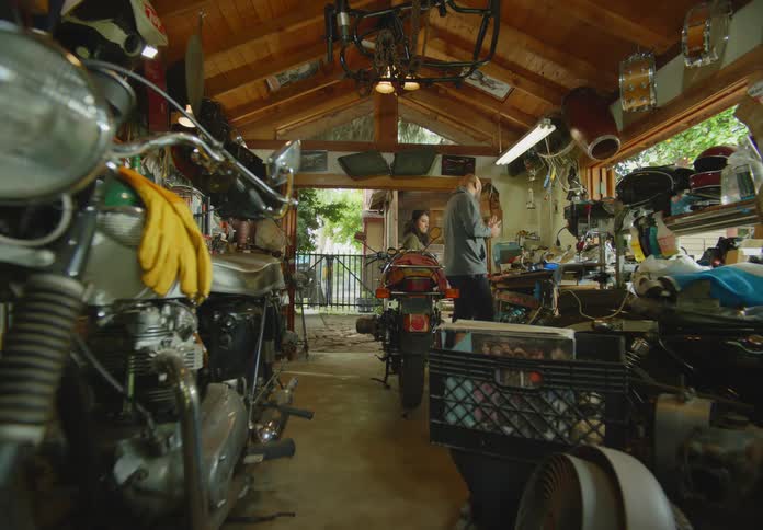 POV from a motorcycle in the foreground of a man and woman in a messy open air garage workshop as they high-five and then the man turns and rubs his hands together and rummages for more parts on the workbench.