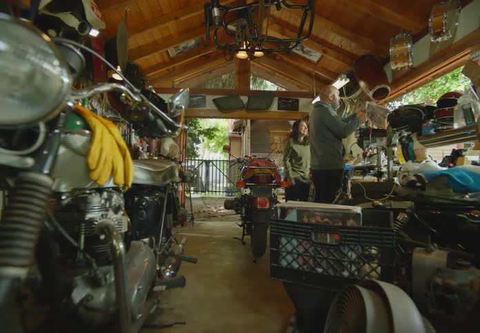 POV from a motorcycle in the foreground of a man and woman in a cluttered open air garage workshop gathering and wiping down parts to work on another motorbike.
