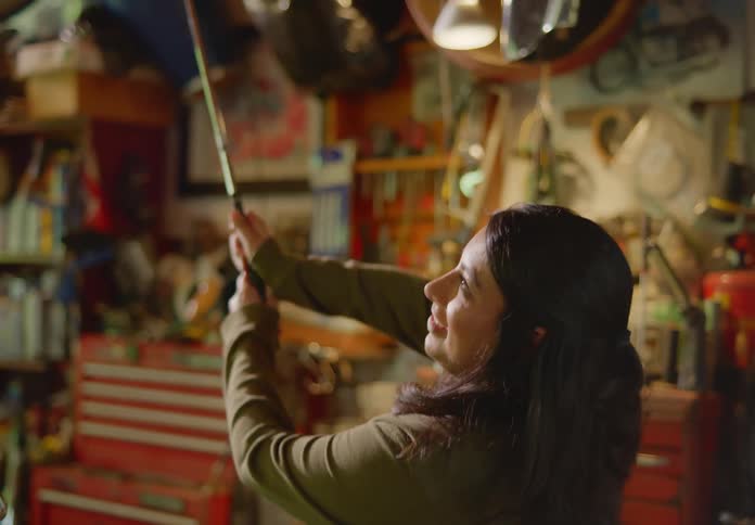 A young brunette woman awkwardly attempts to learn a golf swing with an old set of golf clubs in the garage, then laughs at herself.
