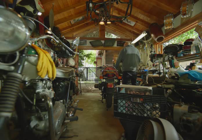 POV from a motorcycle in the foreground of a man and woman in a cluttered open air garage workshop working on another motorcyle.