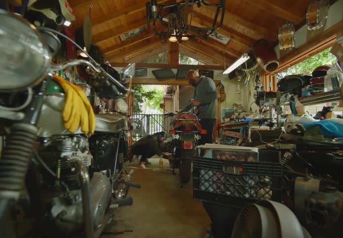 POV from a motorcycle in the foreground of a man and woman in a cluttered open air garage workshop cleaning and wiping down a different motorcyle.