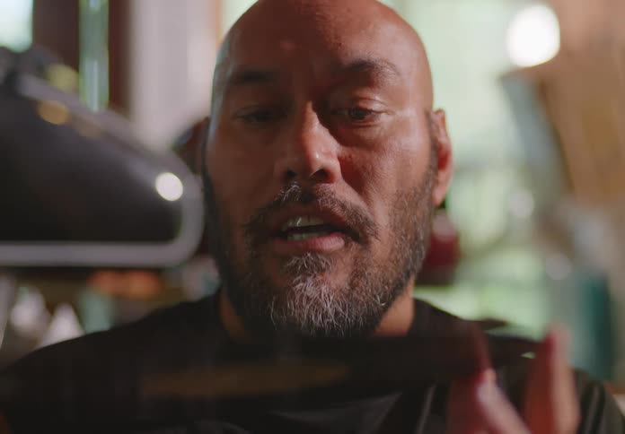 A bald and bearded male Pacific-Islander sits with a young woman in a storage shed and examines an old vinyl record closely as they chat.