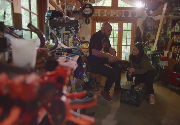 A young couple sit in a cluttered garage surrounded by bikes, tools, and various equipment and go through a crate of old vinyl records together.