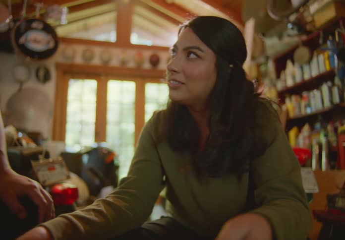 A young dark-haired woman sitting in a messy garage looking through a crate of old vinyl records and laughing as she reminisces with the man sitting beside her.