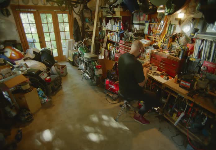 A bald man sitting at a workbench, in a messy garage full of various tools and parts, using a small hammer and chisel to work on a project.