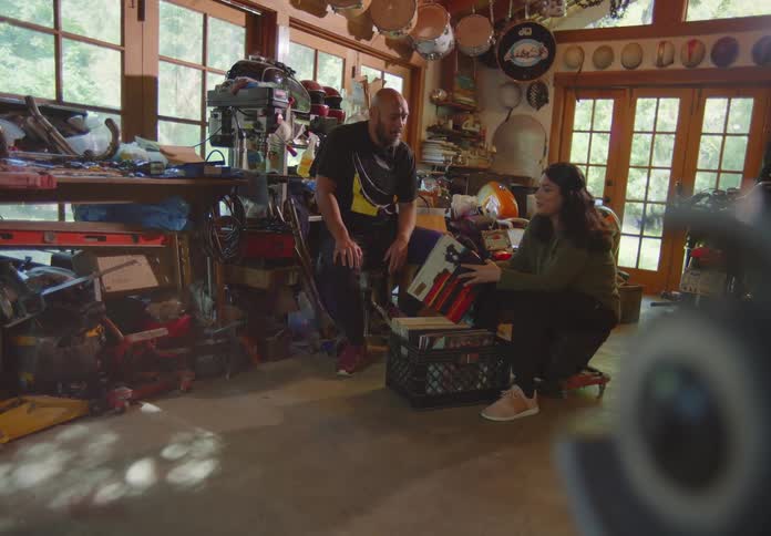 An adult brother and sister look through their parents old vinyl records in a cluttered garage while they reminisce and laugh together.