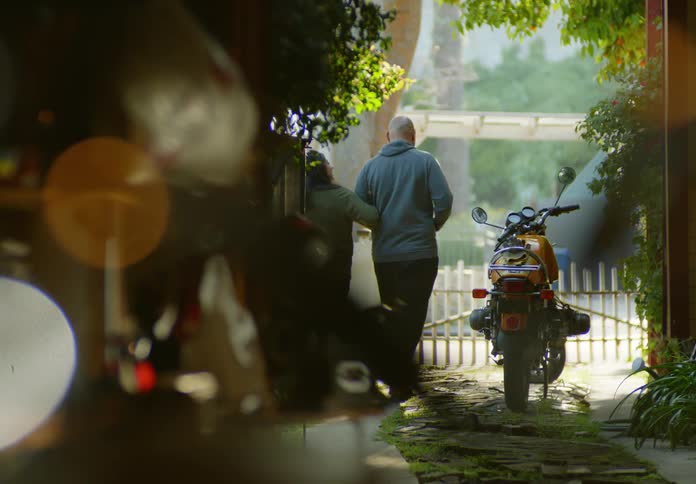 View from behind of a man and woman walking arm in arm at sunset down the driveway and past a parked motorcycle to the front gate and sidewalk where other people are walking.