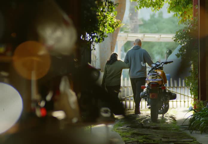 A man and a woman walk arm in arm through a tunnel of trees and past a parked motorcyle in the driveway as he escorts her to the picket fence gate to give her a hug goodbye.