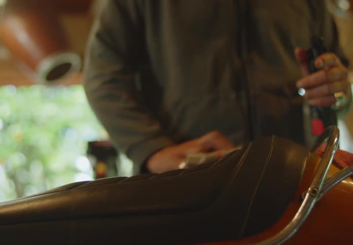A dark, bald, bearded man sprays a rag with a spray bottle and uses the rag to wipe down the seat of a motorbike