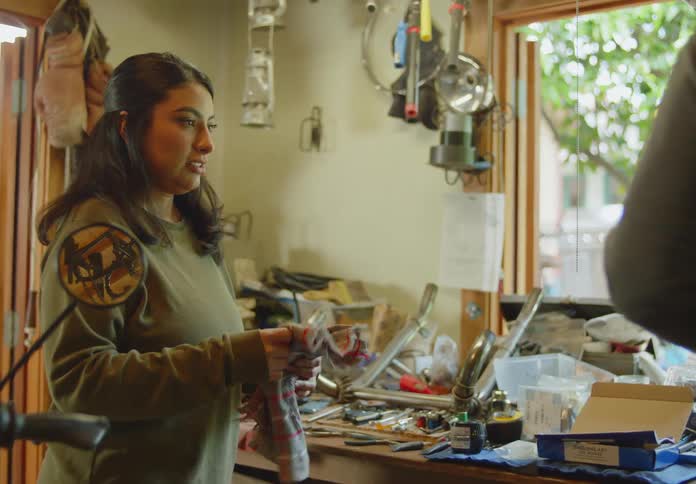 A dark haired woman stands in a messy workshop as a man hands her a rag and spray bottle to clean a motorcycle.
