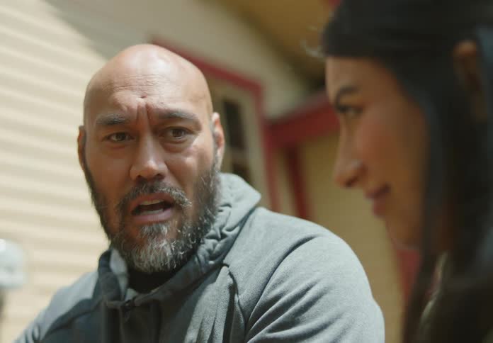 A bald and fit bearded man sitting outdoors furrows his brow and bites his lip as he explains something to a dark-haired female next to him who squints and giggles as they smile and flirt.