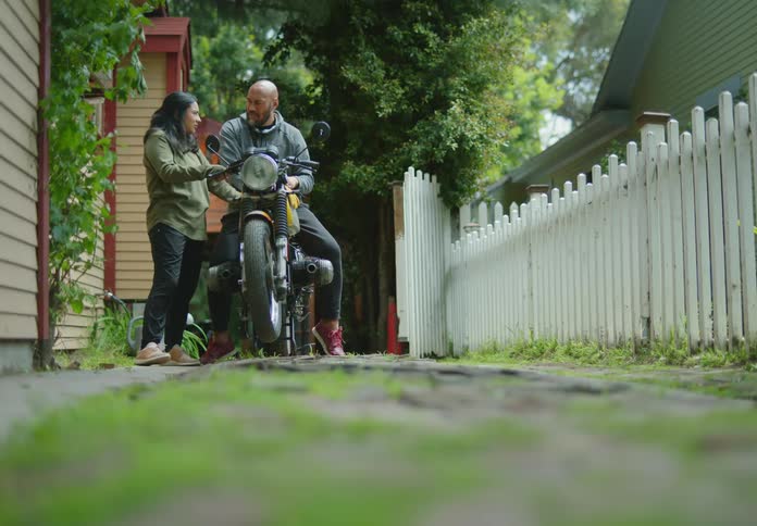 A couple stand in a driveway and examine the motorcycle the man is sitting on while they laugh and talk flirtatiously.