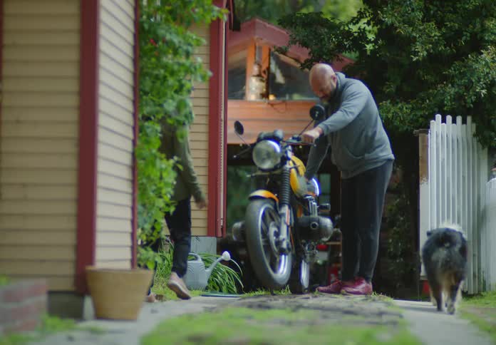 A bald, bearded man pulls out his motorcycle and sits on it while his wife is walking down the gravel driveway toward him with their dogs, and she stops to talk to him.