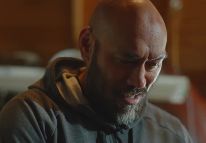 A dark skinned man with a bald head and beard and wearing a hoodie smiles as he applies glue to a part on his motorcycle