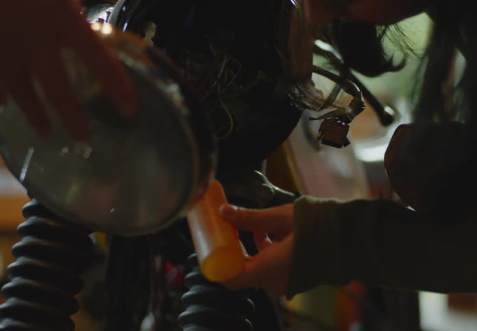 A dark-haired woman applies glue to the light casing on a motorcycle as another person stands by holding the headlight to be installed.