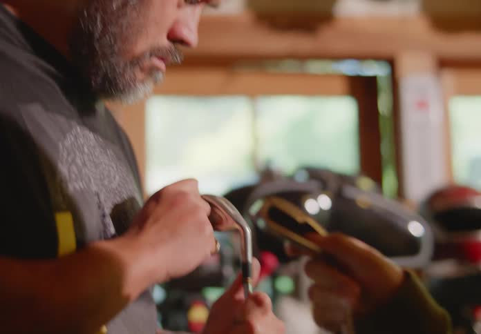 An older bald and bearded man and a young brunette woman hold and examine some golf clubs they found in a cluttered garage.