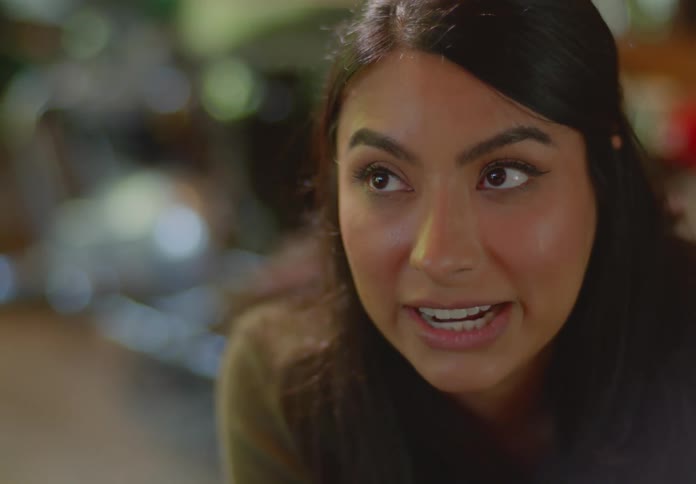 A seated young, ethnic woman looking up from under her eyebrows, listens and responds to someone offscreen.